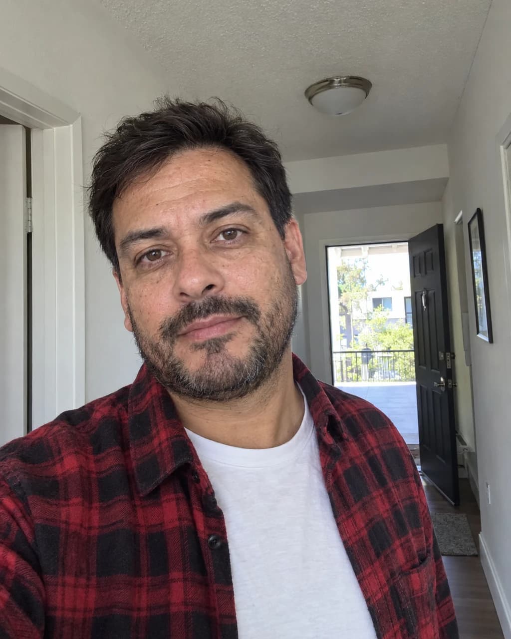 Example person 6: a selfie of a Latino man in his forties with a short beard, in a red flannel shirt in an apartment hallway.